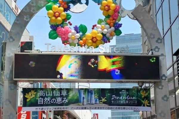 Une rue ornée d’une grande arche décorée de ballons multicolores, de diverses enseignes de magasins et d’un panneau publicitaire numérique.