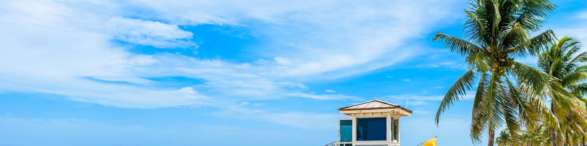 A beach with a lifeguard tower, palm trees, and people relaxing under umbrellas.