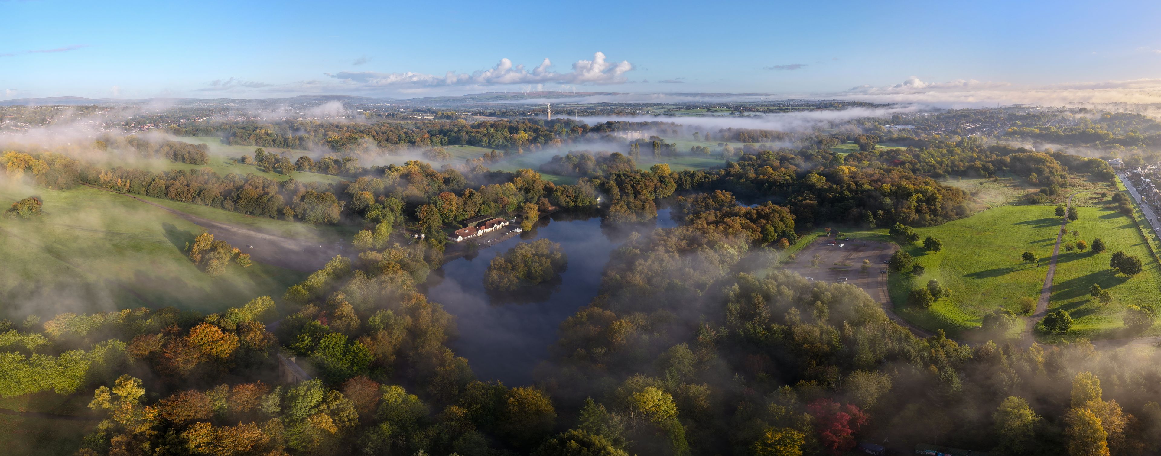 A misty landscape with a river, green fields, and scattered trees.