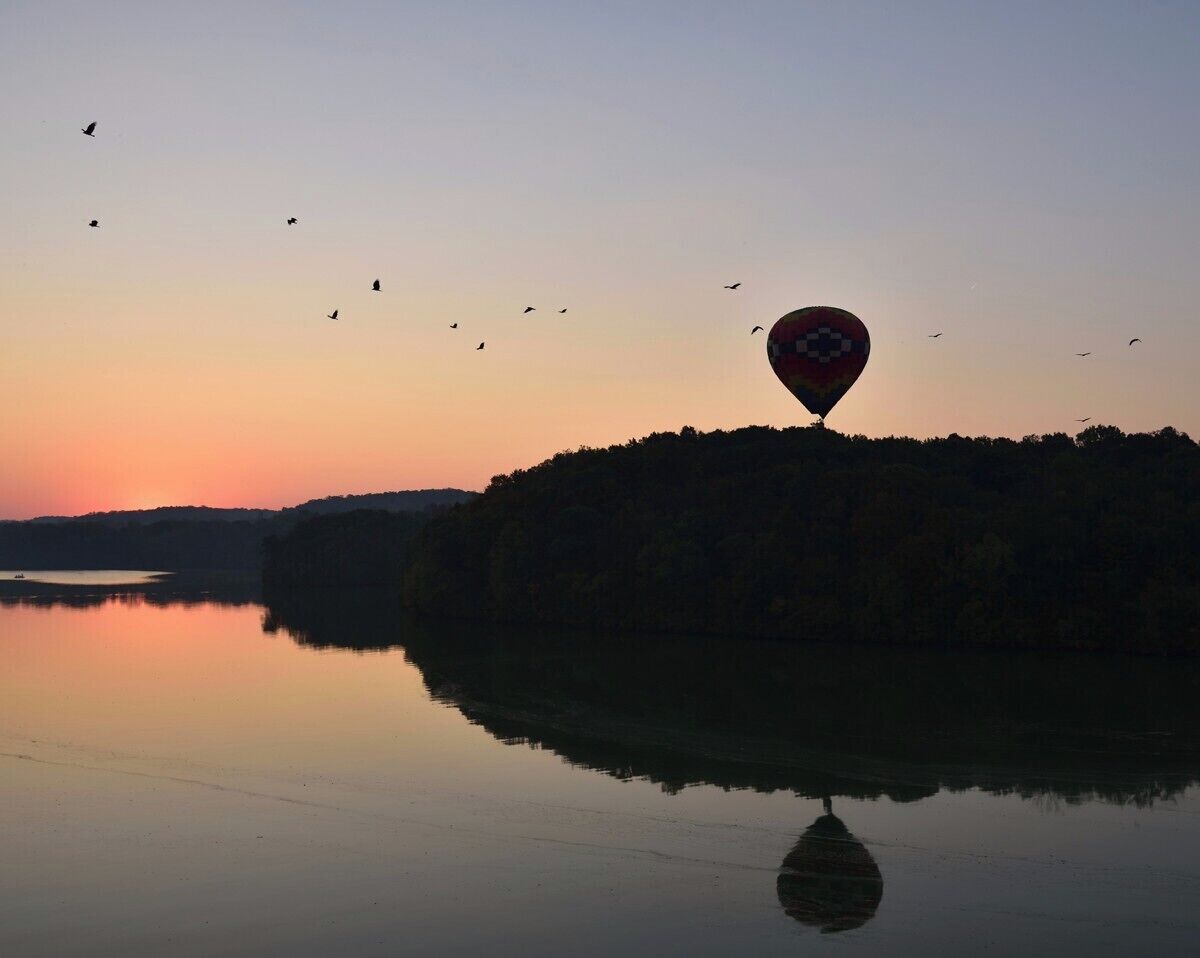 A hot air balloon over a calm lake at sunset with birds flying around.