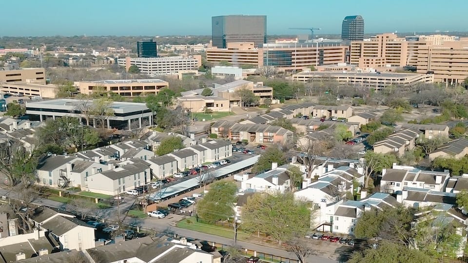 Panorama aerial dense urban housing near Fair Oaks Ave and Holly Hill Dr reveals tightly packed apartments leading toward commercial towers horizon. Brown roofs, parked cars, midtown Dallas, TX