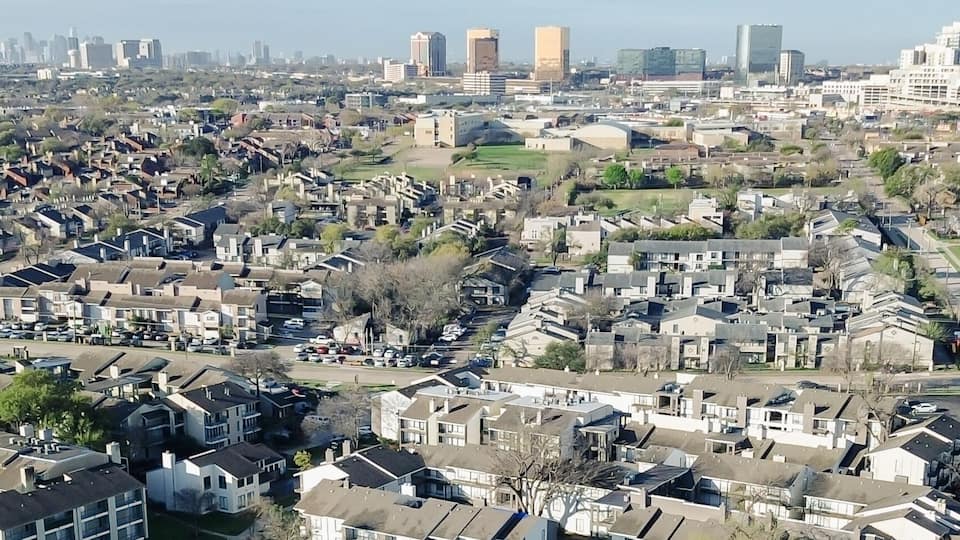 Panorama aerial urban edge overlook near Fair Oaks Ave and Walnut Hill Ln reveals apartments bordering wooded trails with skyline rising beyond. Sloped roofs, mixed facades, cool winter, Dallas