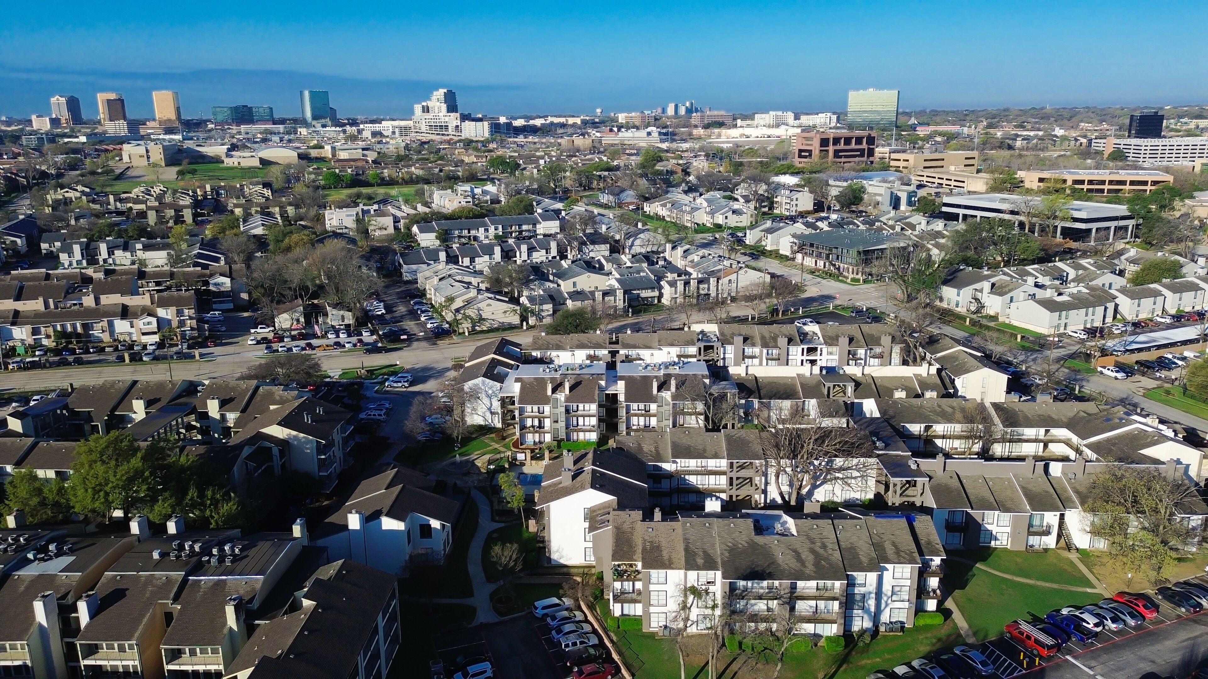 Multifamily–skyline approach near Fair Oaks Ave, Pineland Dr highlights residential clusters extending toward rising office buildings. Light exteriors, patterned roofs, muted foliage rest, Dallas