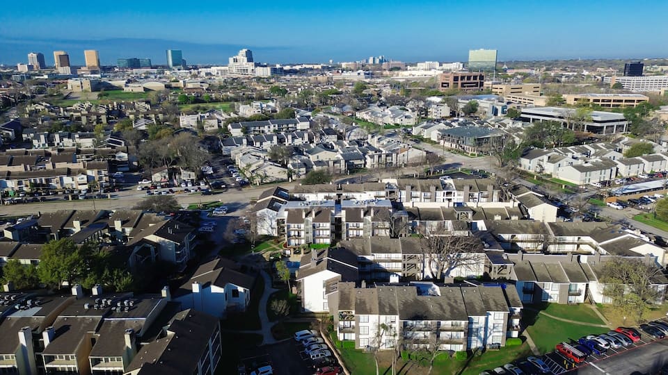 Multifamily–skyline approach near Fair Oaks Ave, Pineland Dr highlights residential clusters extending toward rising office buildings. Light exteriors, patterned roofs, muted foliage rest, Dallas