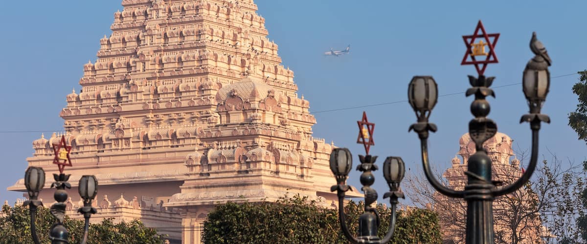 View of lamps with a traditional Hindu six-pointed star, and the main building of the Chattarpura temple complex near by which the passenger plane flies by.