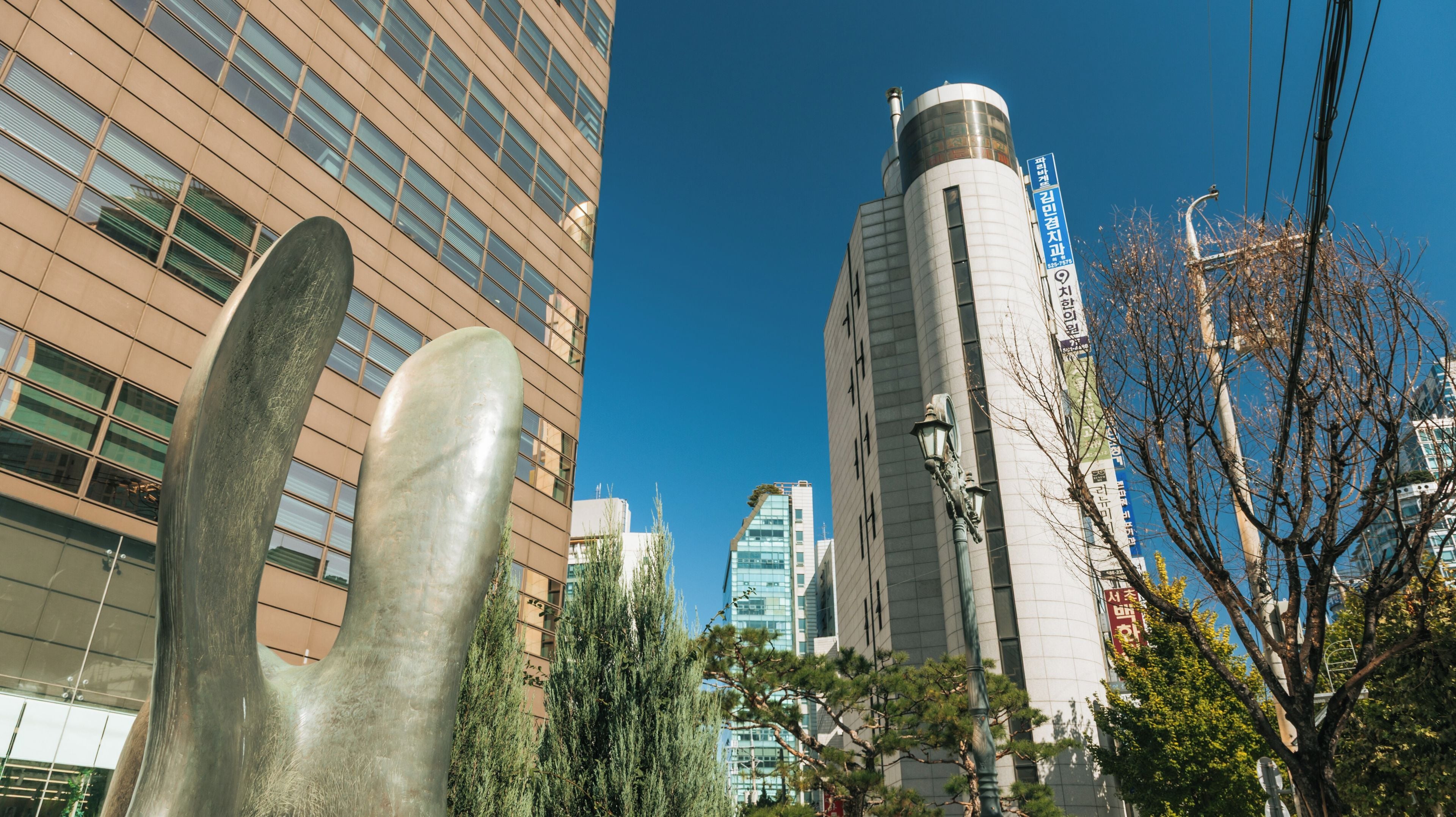A modern urban landscape with a tall cylindrical building, a large metallic sculpture, and a mix of modern and traditional architecture.