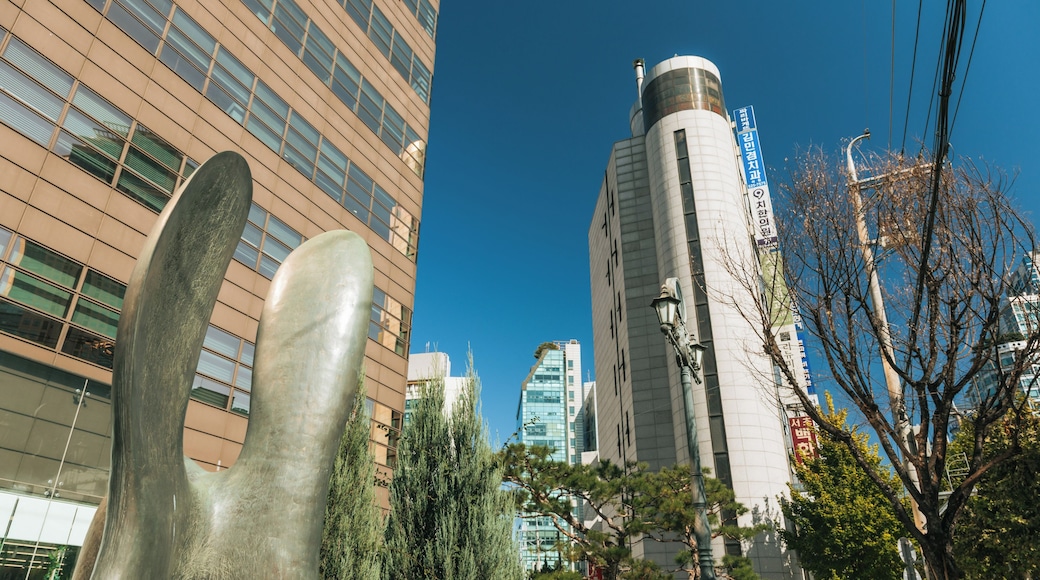 A modern urban landscape with a tall cylindrical building, a large metallic sculpture, and a mix of modern and traditional architecture.