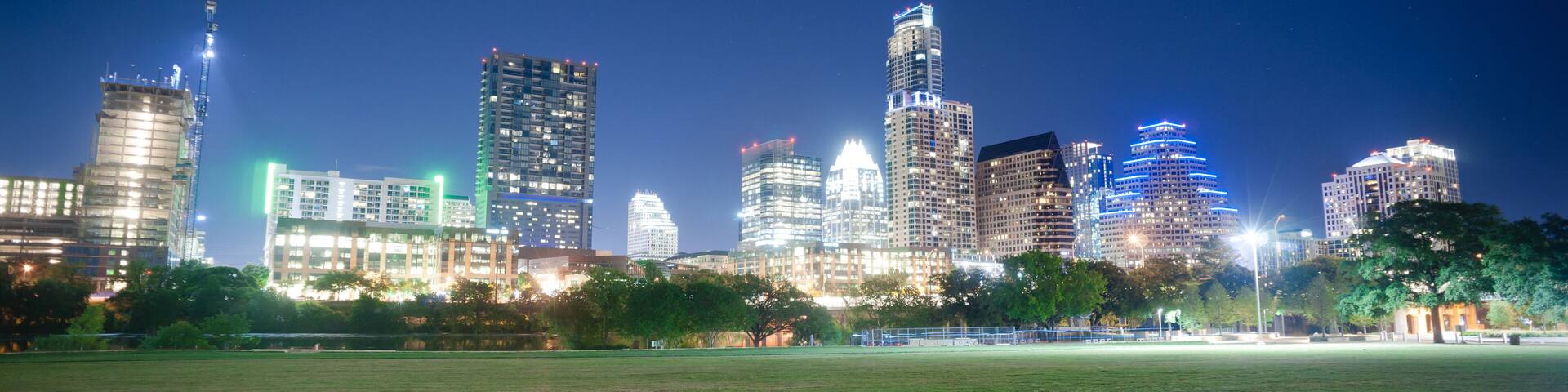 A city skyline at night with illuminated buildings and a large open grassy field in the foreground.