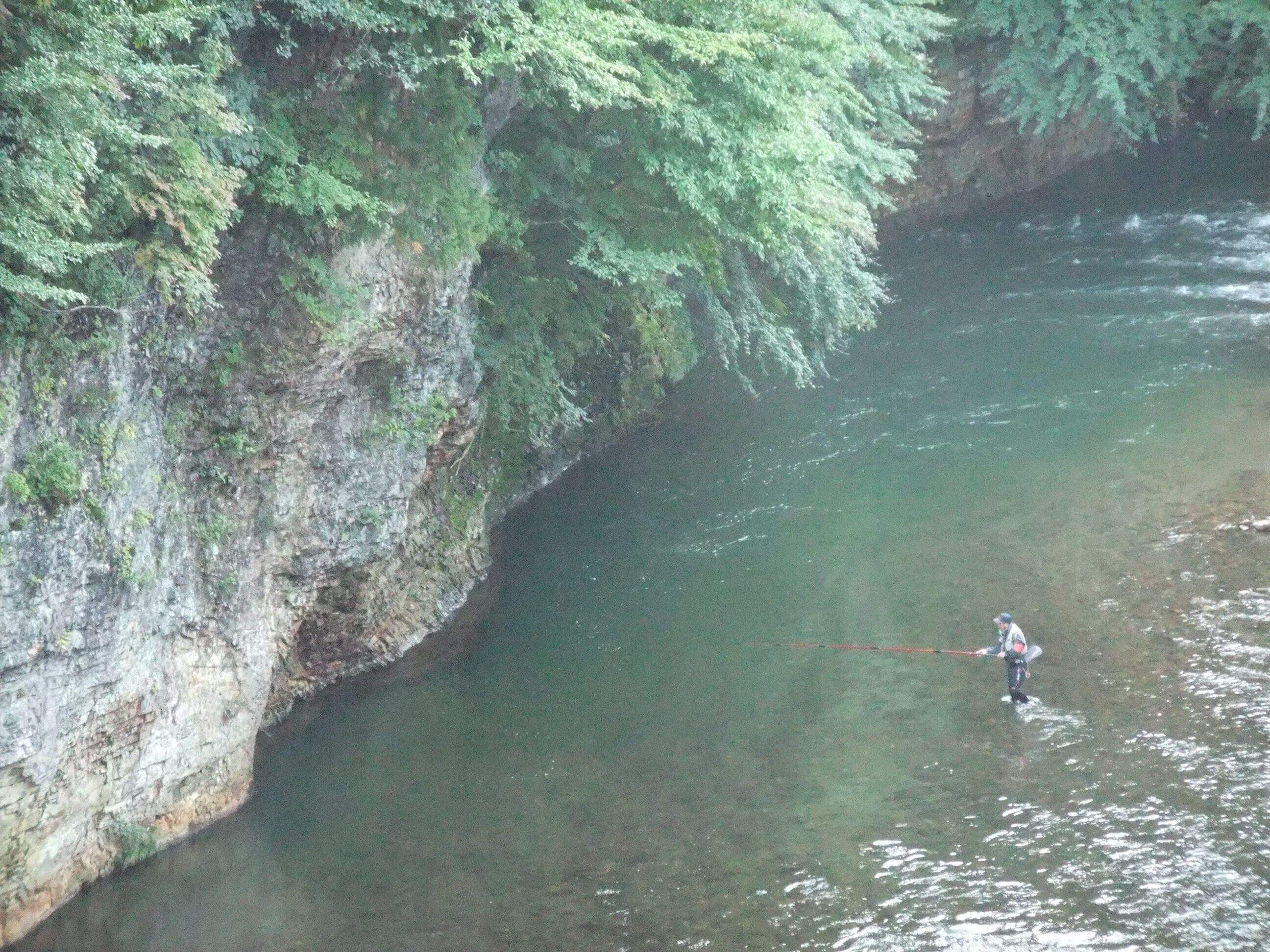 A person fishing in a river with a rocky cliff on one side.