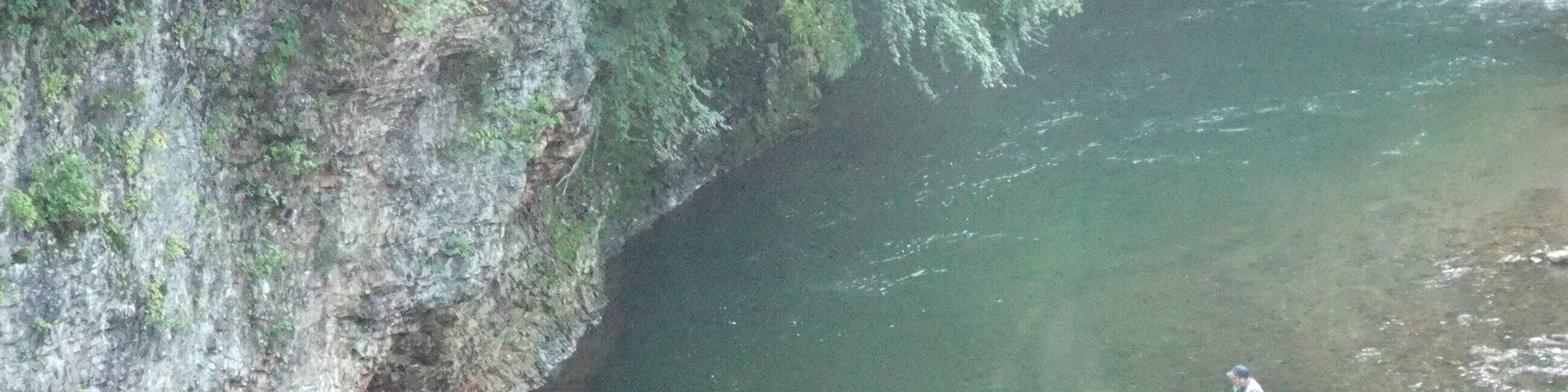 A person fishing in a river with a rocky cliff on one side.