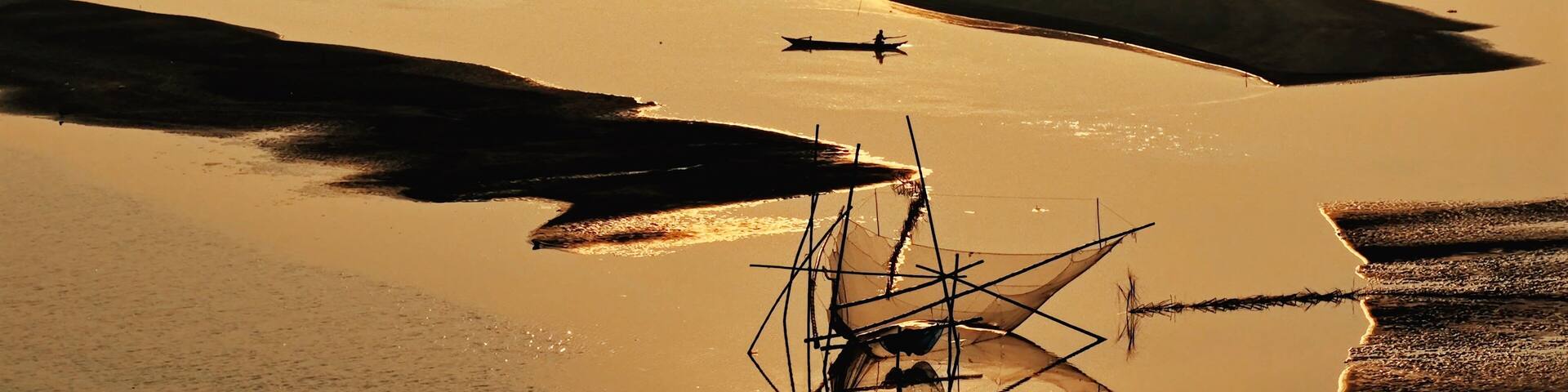 A traditional fishing net stands silhouetted against the golden waters of the Brahmaputra River at sunset in Majuli, Assam, as sandbars emerge during the dry season