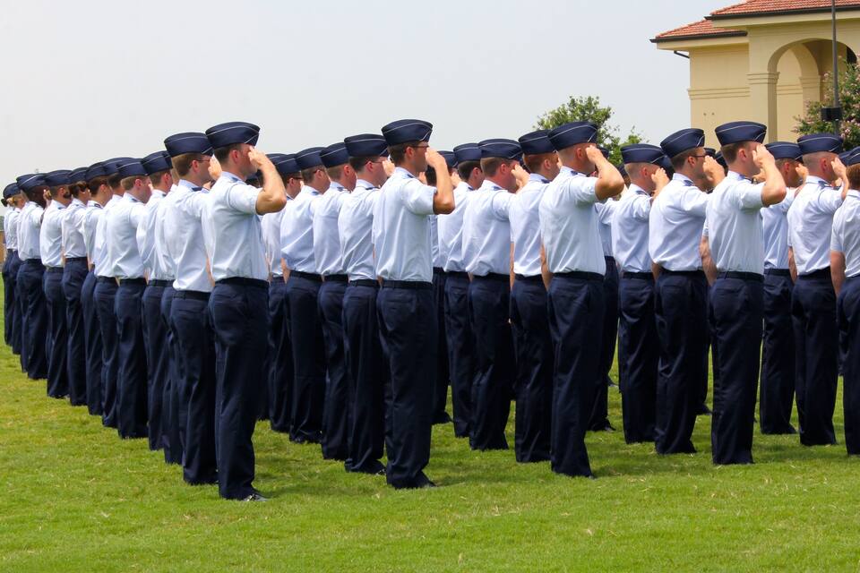 A group of uniformed individuals in white shirts and dark trousers saluting in formation on a grassy field with a building in the background.