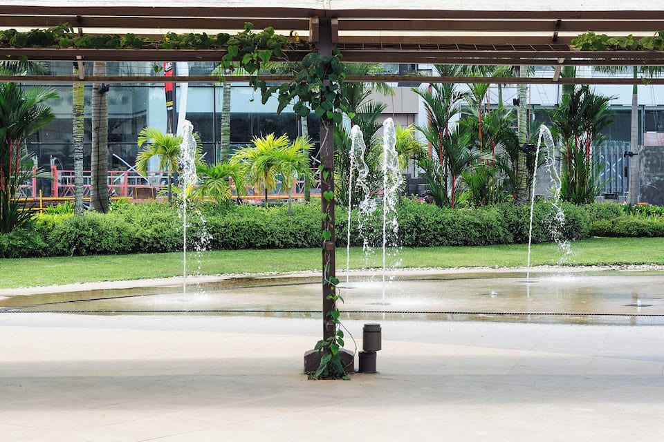 Una fuente con chorros de agua en un parque con palmeras y un edificio al fondo.