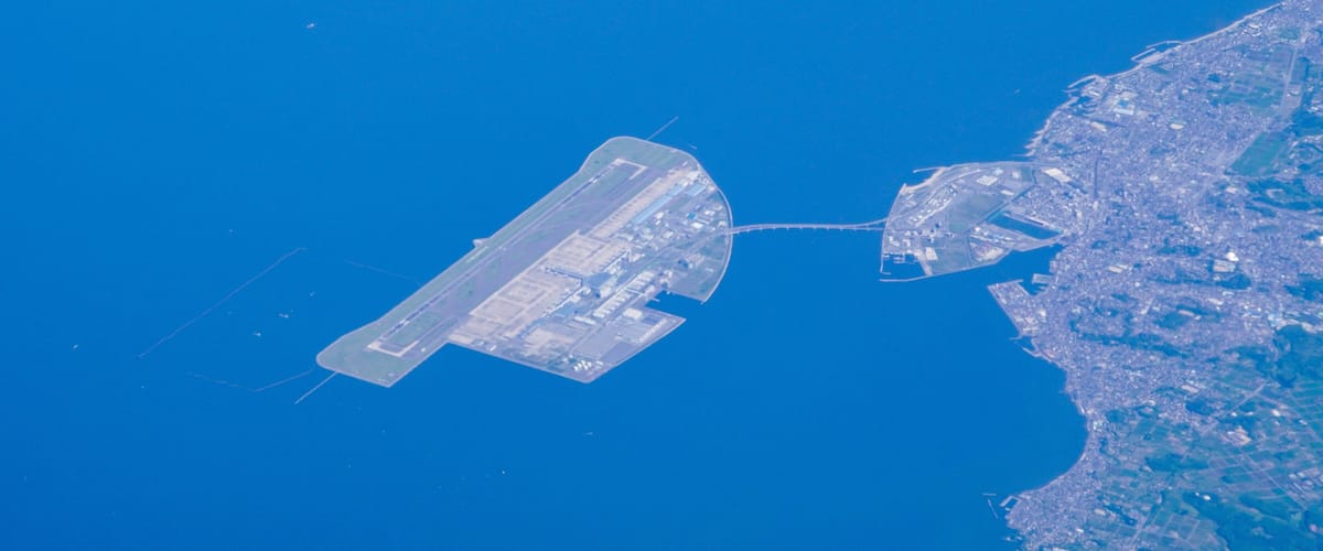 An aircraft carrier and a smaller ship docked at a port near a coastline.