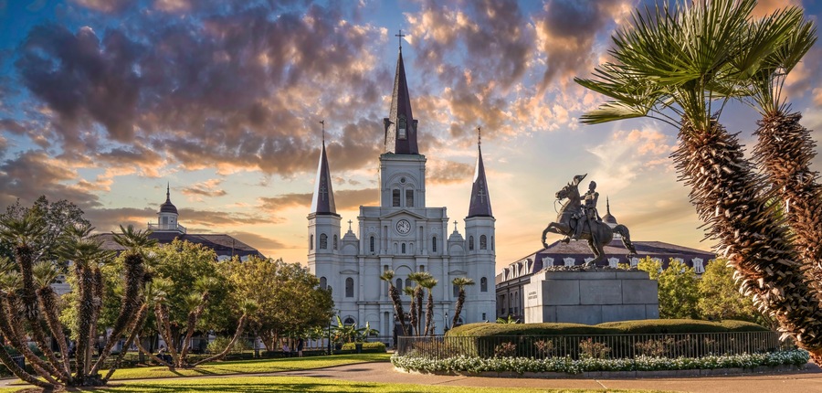 A historic church with two tall spires, a statue of a mounted figure, and lush greenery.