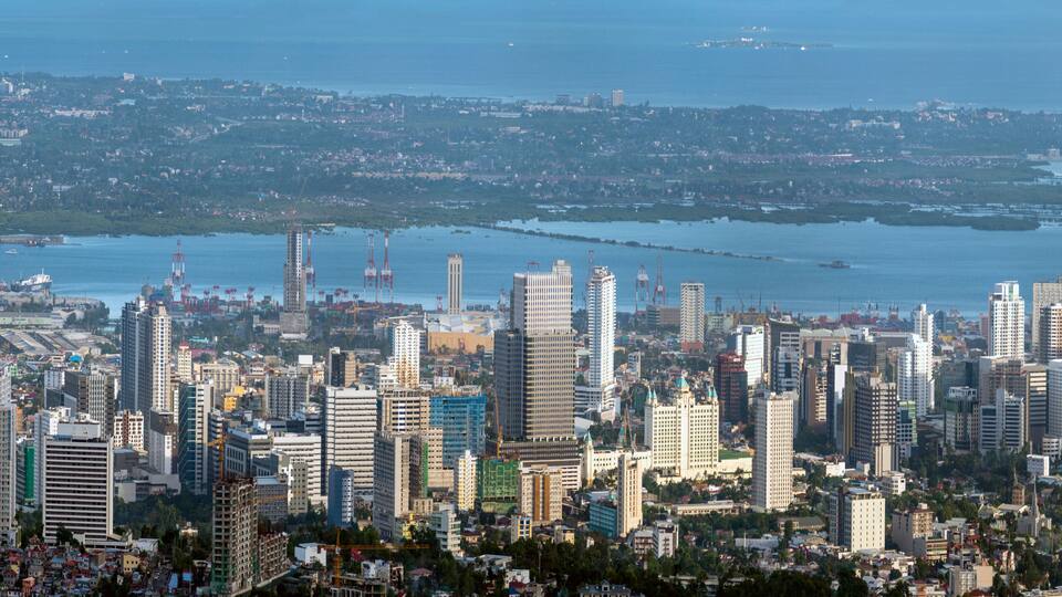 A cityscape with numerous high-rise buildings, a waterfront, and a distant view of a hilly landscape.