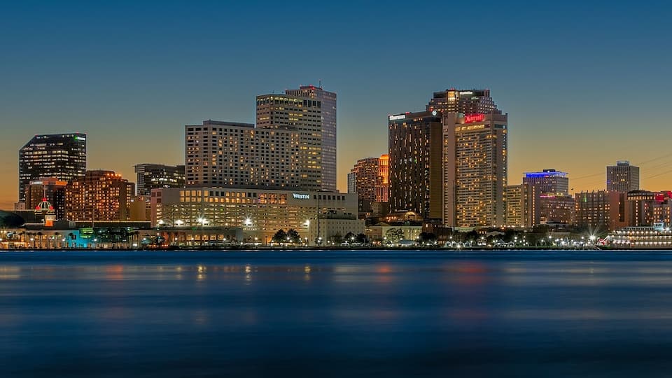 A city skyline at dusk with illuminated buildings and a calm river in the foreground.