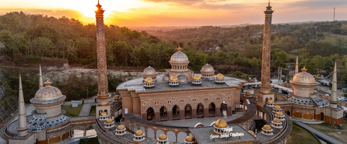 Aerial view of Masjid Samin Baabul Muttaqien with its intricate golden domes and tall minarets surrounded by lush greenery during a warm sunset in Bojonegoro, East Java, Indonesia.