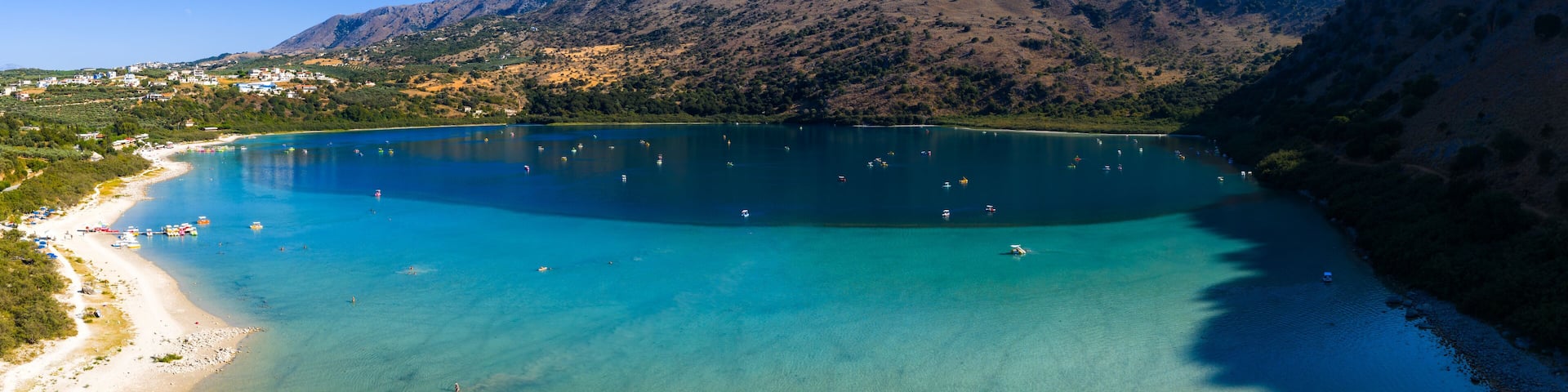 Aerial view of Lake Kournas in Crete, Greece, featuring turquoise waters, boats, a sandy shoreline with umbrellas, and surrounding rugged hills.