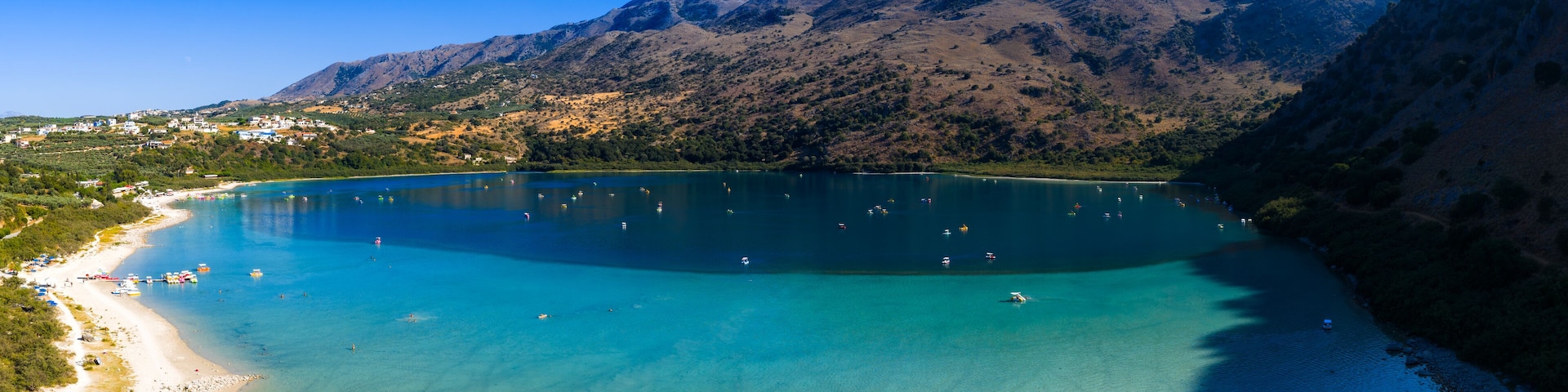 Aerial view of Lake Kournas in Crete, Greece, featuring turquoise waters, boats, a sandy shoreline with umbrellas, and surrounding rugged hills.