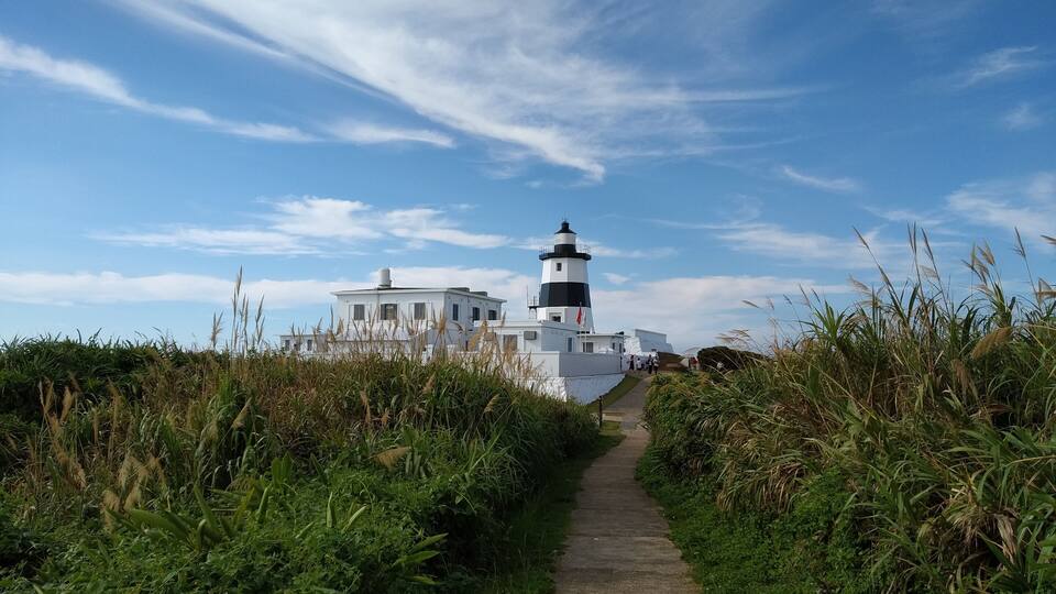 A lighthouse with a white building in the foreground and a clear blue sky with wispy clouds.