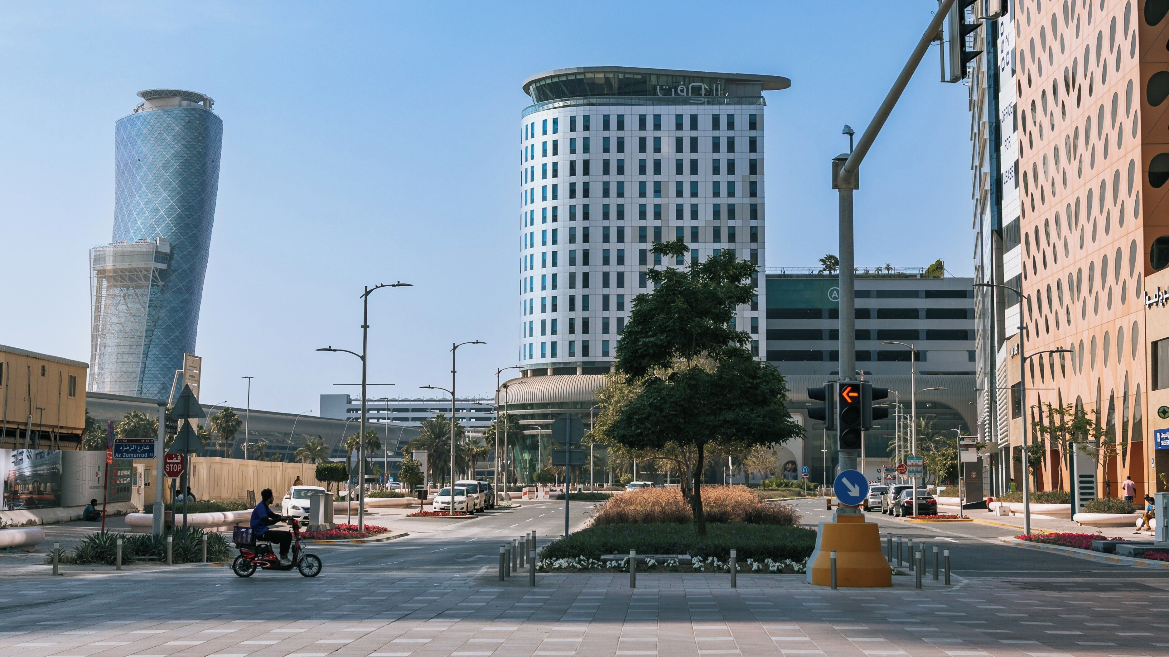 Cruzamento urbano com edifícios modernos, uma placa de pare e um semáforo, e uma pessoa andando de bicicleta.