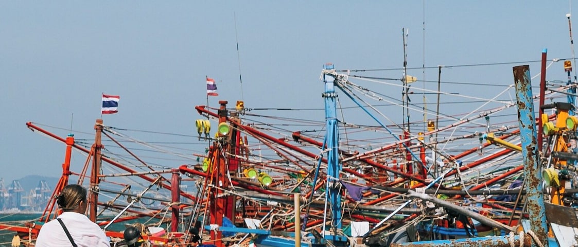 Um movimentado porto com muitos barcos de pesca, uma mulher caminhando e diversos equipamentos de pesca espalhados pelo chão.