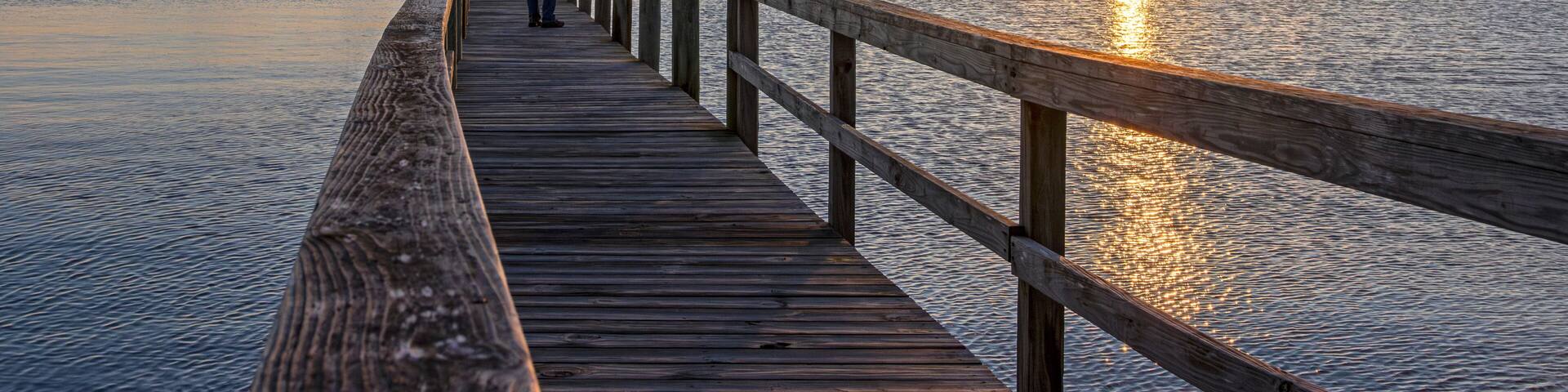 Man Standing At End Of Dock On A Bay At Sunrise Time