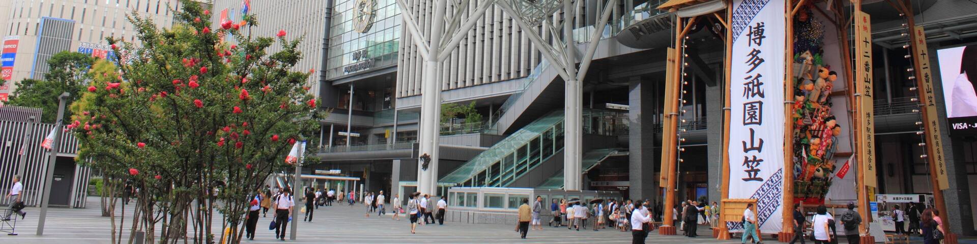 A modern building with a distinctive curved glass facade and a large open plaza with trees and people.
