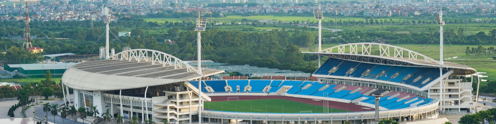 Ein großes Stadion mit blauen Sitzplätzen und einem grünen Rasen, umgeben von Grünflächen und einer Stadtkulisse im Hintergrund.