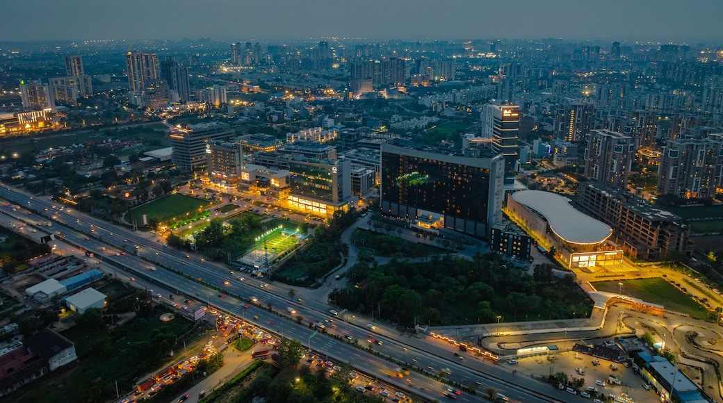Aerial view of the modern cityscape at dusk, featuring illuminated skyscrapers, office buildings, and a busy highway with light trails from traffic in Gurgaon, Haryana, India.