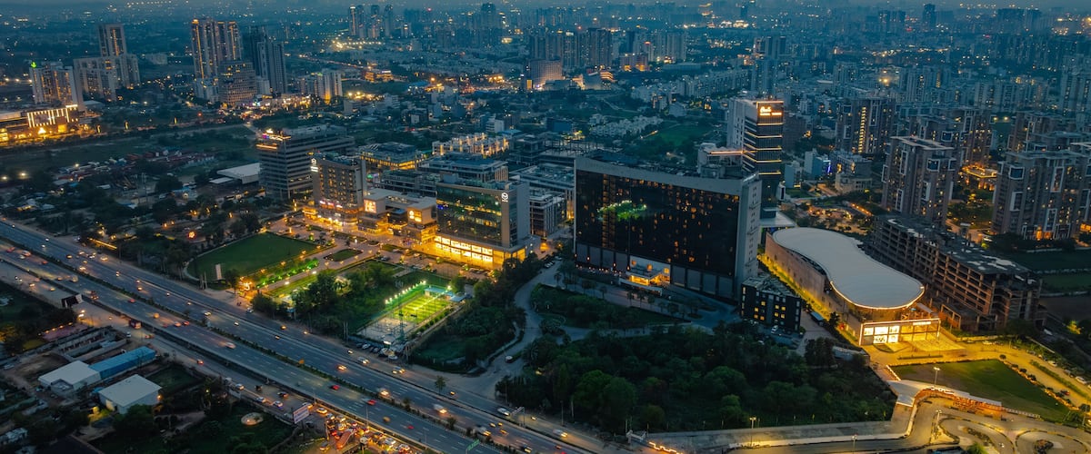 Aerial view of the modern cityscape at dusk, featuring illuminated skyscrapers, office buildings, and a busy highway with light trails from traffic in Gurgaon, Haryana, India.