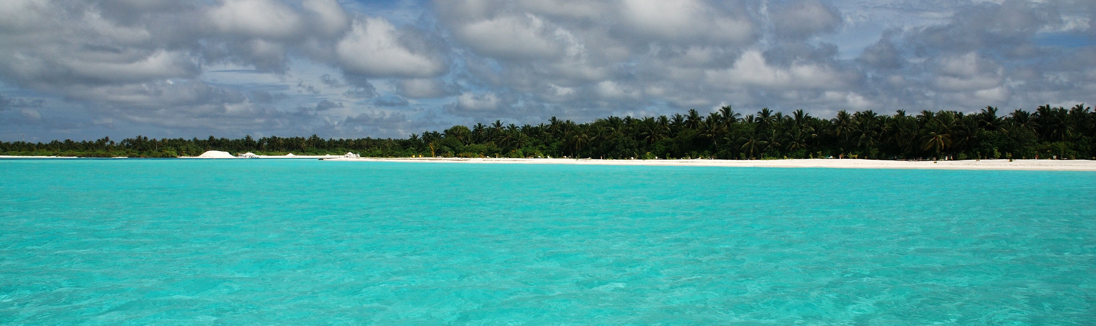 Turquoise waters, a sandy beach, and a line of palm trees under a cloudy sky.