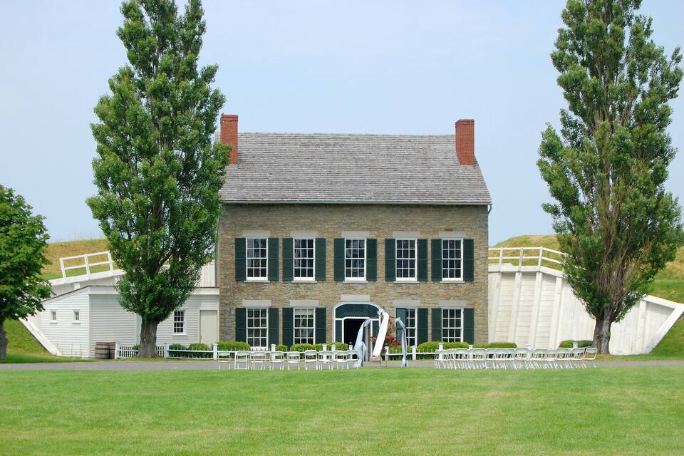 A historic stone building with green shutters and a central entrance, surrounded by a grassy lawn and two tall trees.