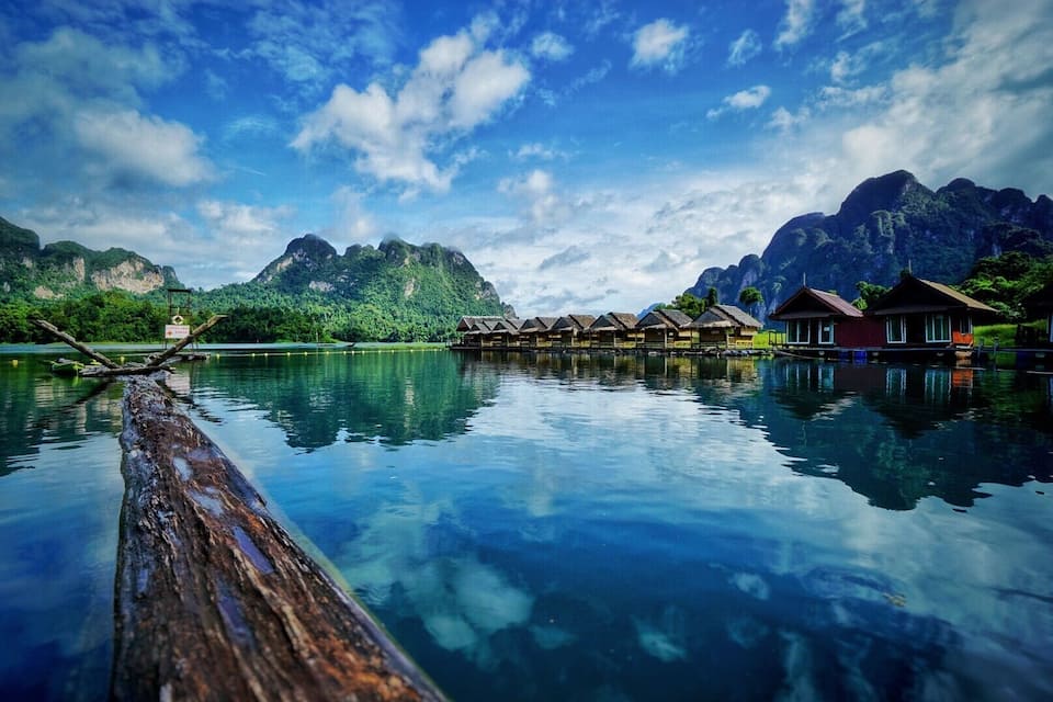 Ein ruhiger See mit Holzhäusern auf Stelzen, umgeben von üppigen grünen Bergen und blauem Himmel.