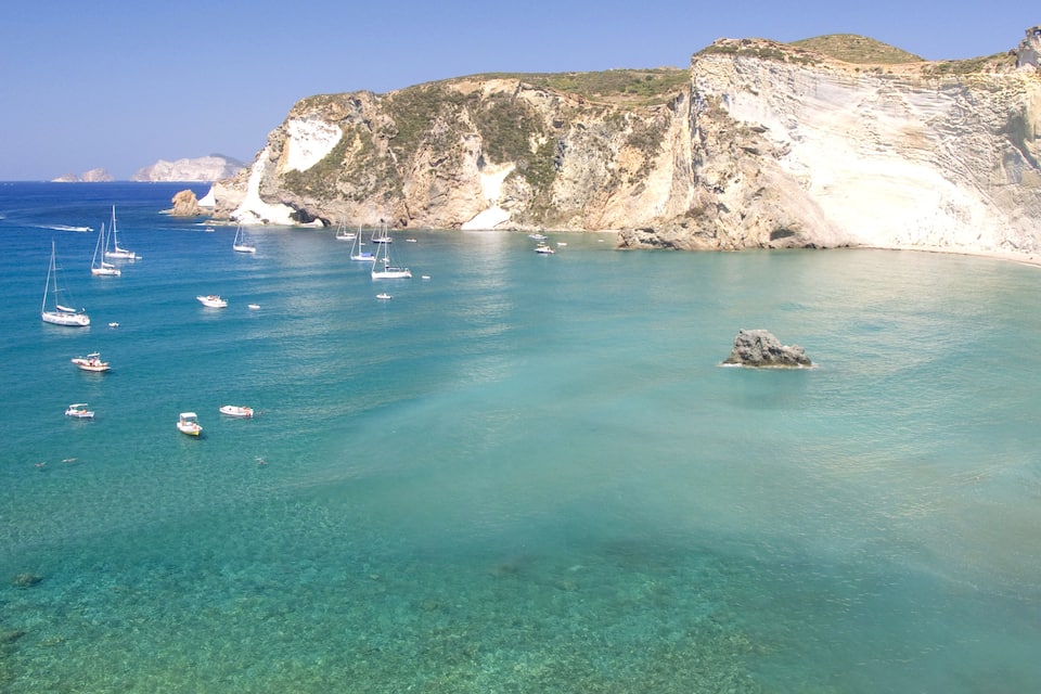 A coastal area with clear turquoise waters, several sailboats, and rocky cliffs.
