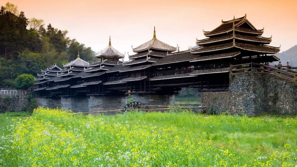 A traditional multi-tiered pagoda with a stone base, surrounded by lush greenery and a field of yellow flowers.