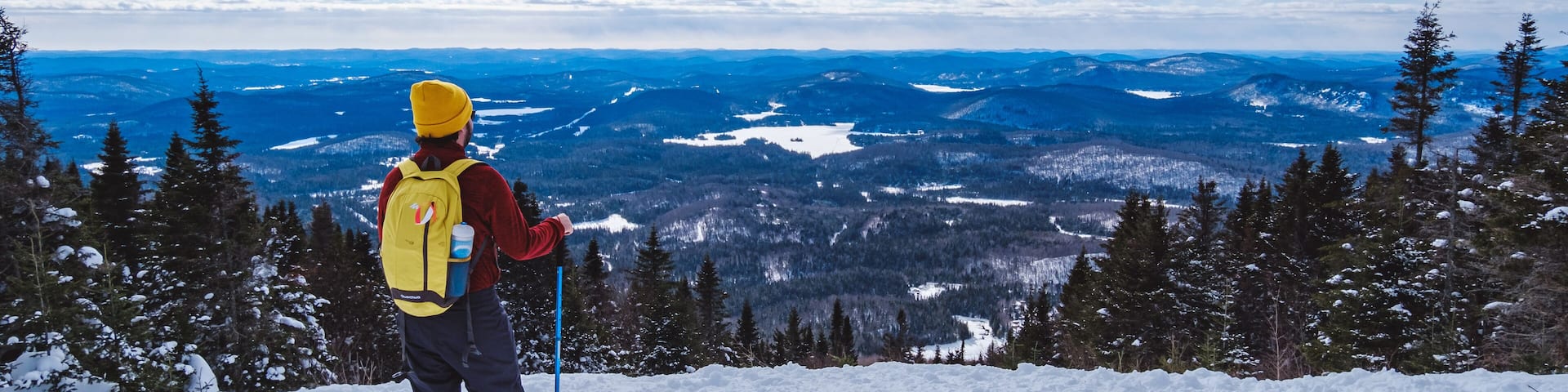 Man with yellow beanie and and yellow backpack looking at the the view from the top of Mont Kaaikop in winter in Quebec (Canada)