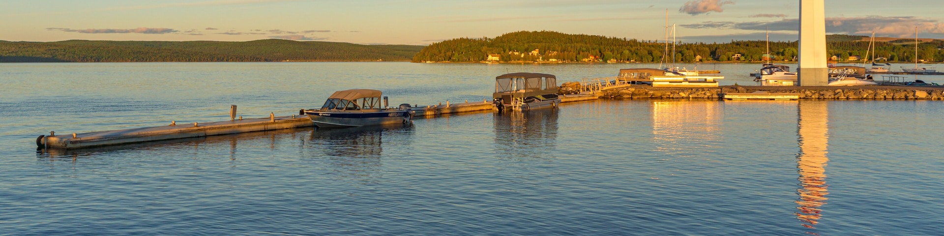 Un lac paisible où des bateaux sont amarrés à un quai, un phare et, au loin, une colline boisée.