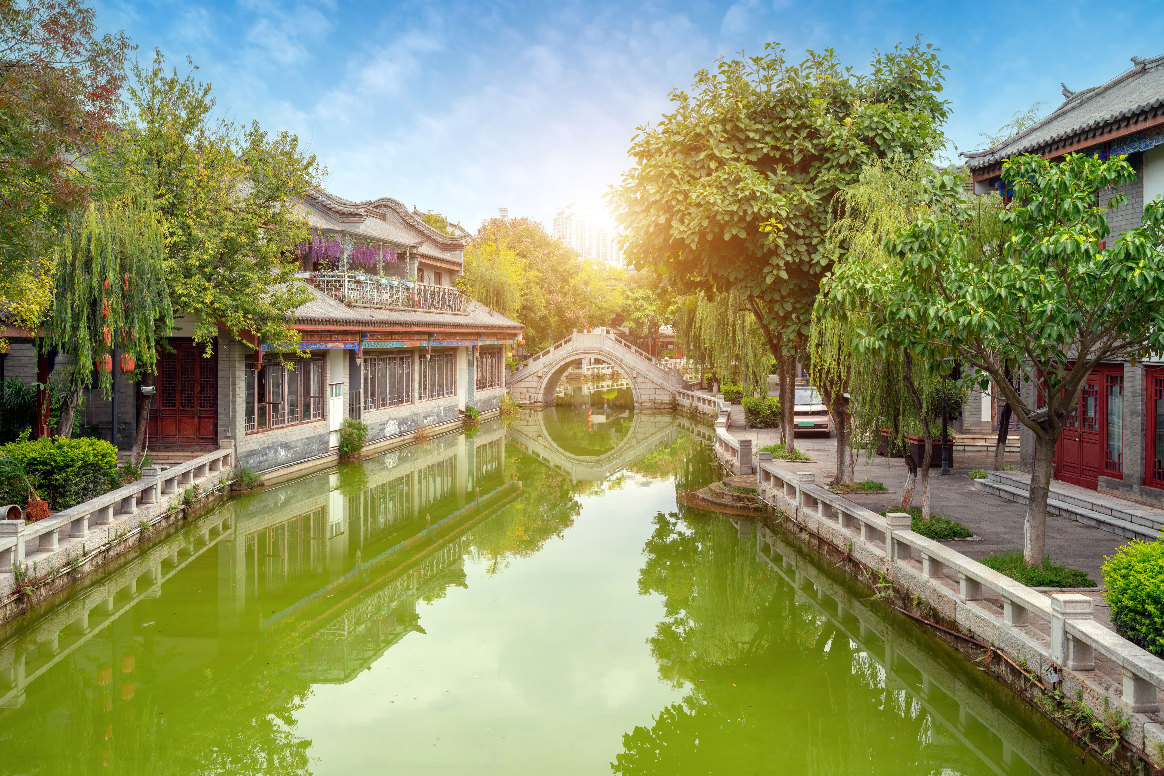 A traditional Chinese waterway with a stone bridge, surrounded by lush greenery and traditional buildings.