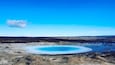A geothermal area with a large blue pool surrounded by rocky terrain and distant hills.
