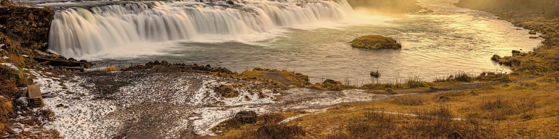 A waterfall cascading into a river surrounded by a snowy landscape with a sunset in the background.