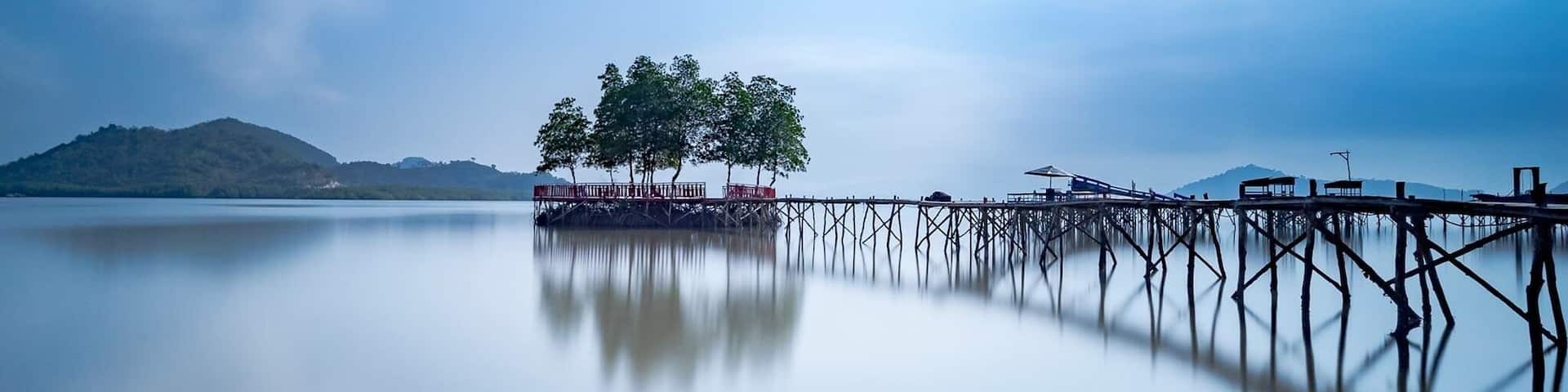 A calm lake with a wooden pier extending into the water, surrounded by trees and a distant mountain range.