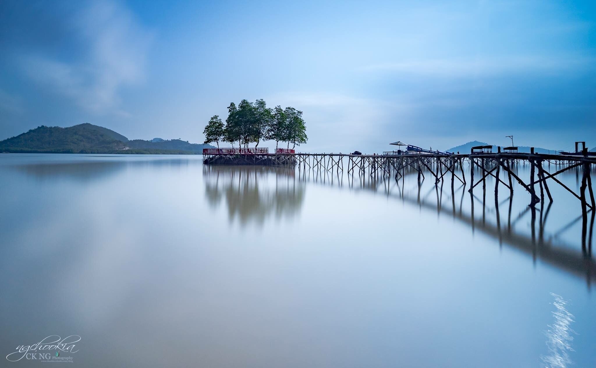 A calm lake with a wooden pier extending into the water, surrounded by trees and a distant mountain range.