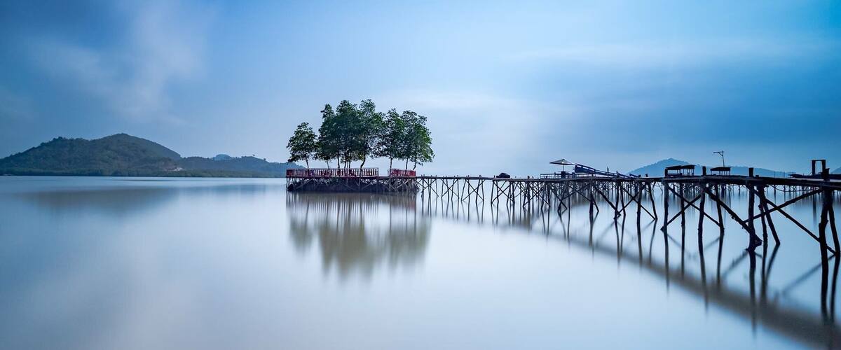 A calm lake with a wooden pier extending into the water, surrounded by trees and a distant mountain range.