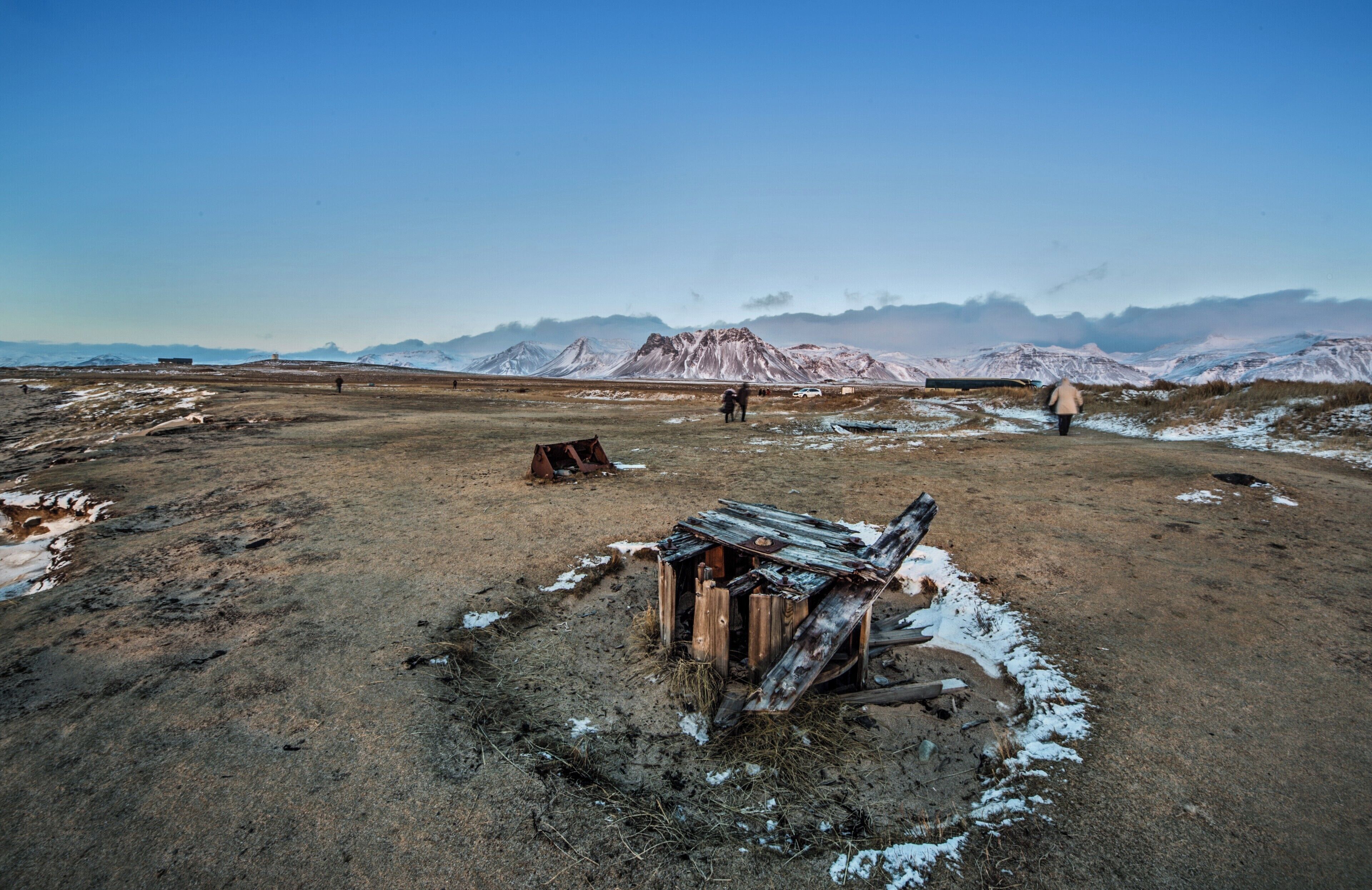 A barren landscape with snow patches, a dilapidated wooden structure, and mountains in the background.