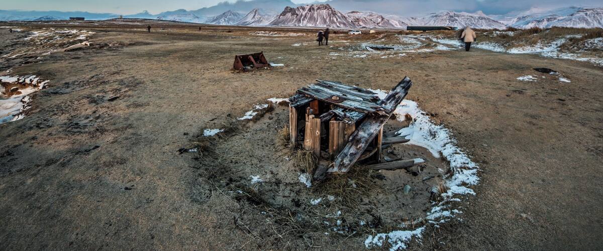 A barren landscape with snow patches, a dilapidated wooden structure, and mountains in the background.