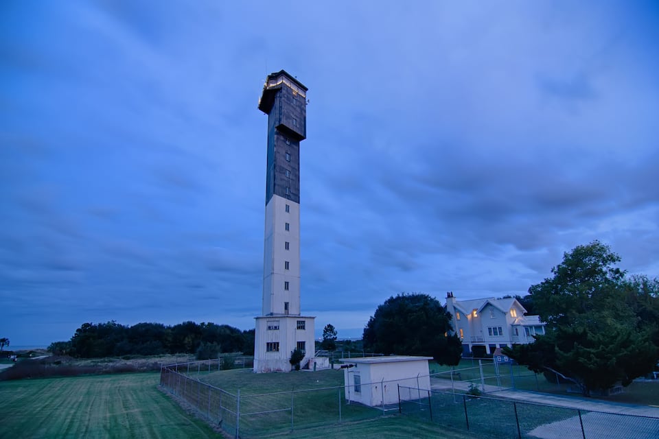 Una torre alta con base blanca y parte superior oscura, rodeada de un área verde y una casa con ventanas iluminadas.