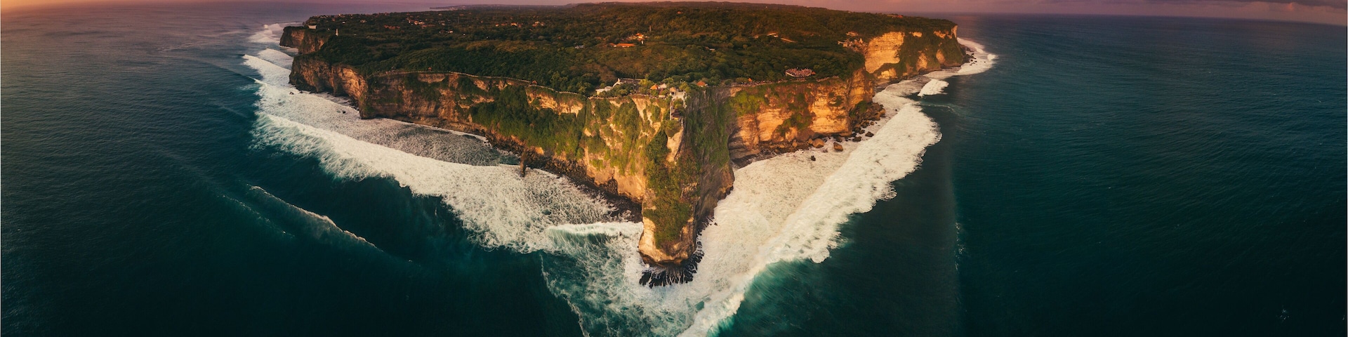 As the sun sets, a drone captures Greece's cliffs, their edges glowing gold, descending into the tranquil turquoise abyss below.