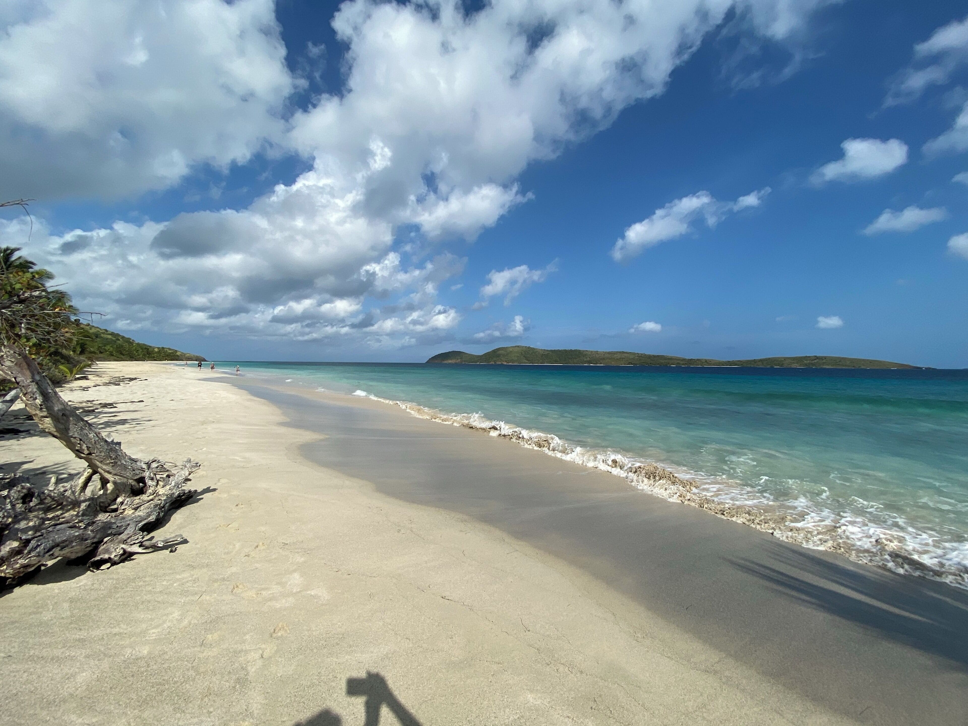 A beach with clear turquoise water, a sandy shore, and distant green hills under a partly cloudy sky.
